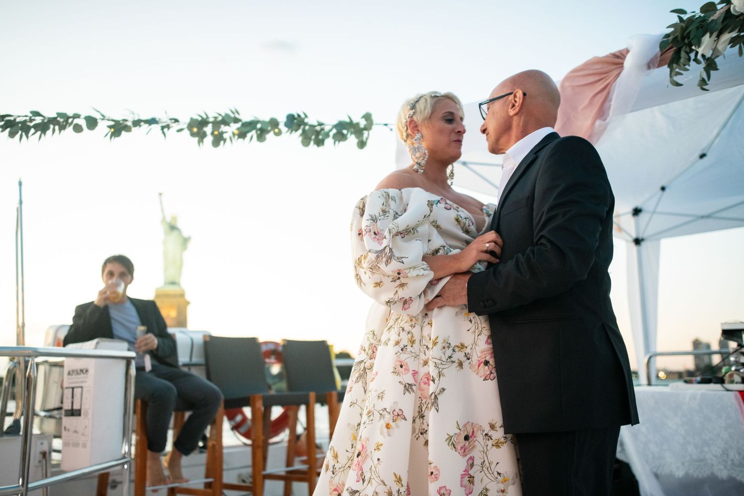 A couple shares a romantic dance moment under floral decorations at an evening waterfront reception venue.