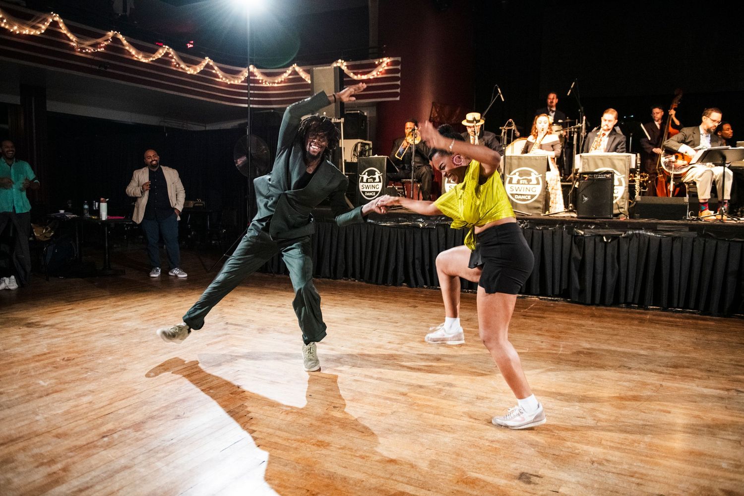 Dancers perform high-energy swing moves on wooden floor with big band in background.