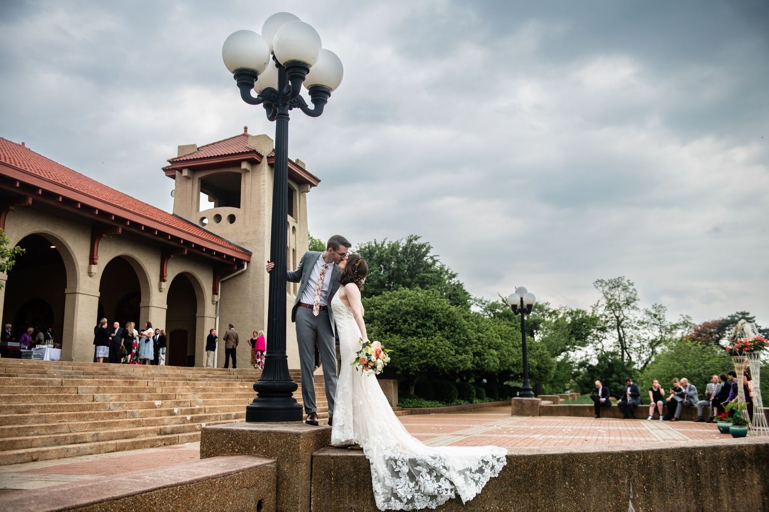 Dramatic wedding photo on steps of historic venue with ornate lamp post and cloudy sky.