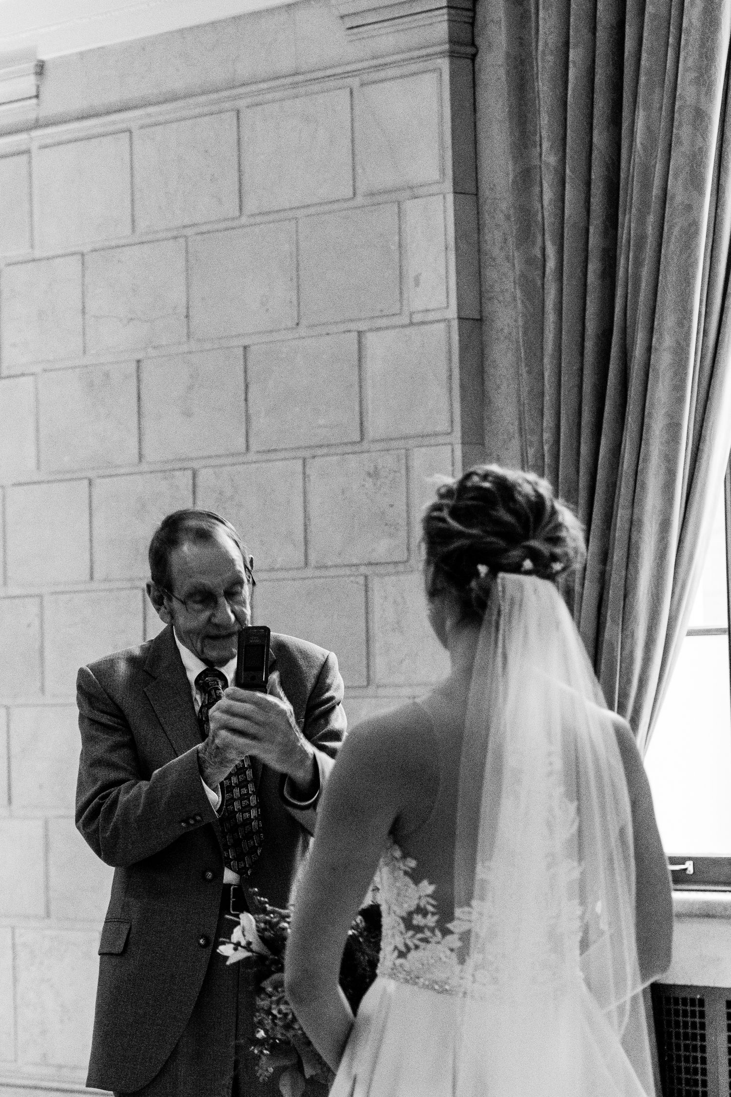 Black and white photo of a father looking at his phone during a wedding ceremony.
