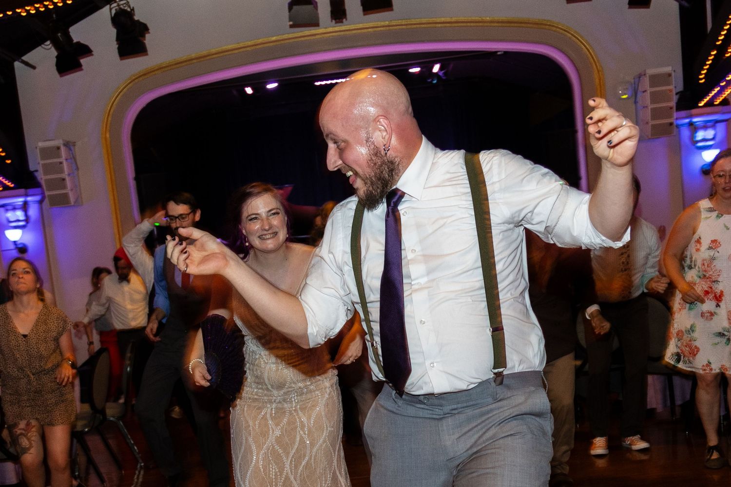 Couple dancing energetically at a wedding reception in an ornately decorated venue with purple lighting.
