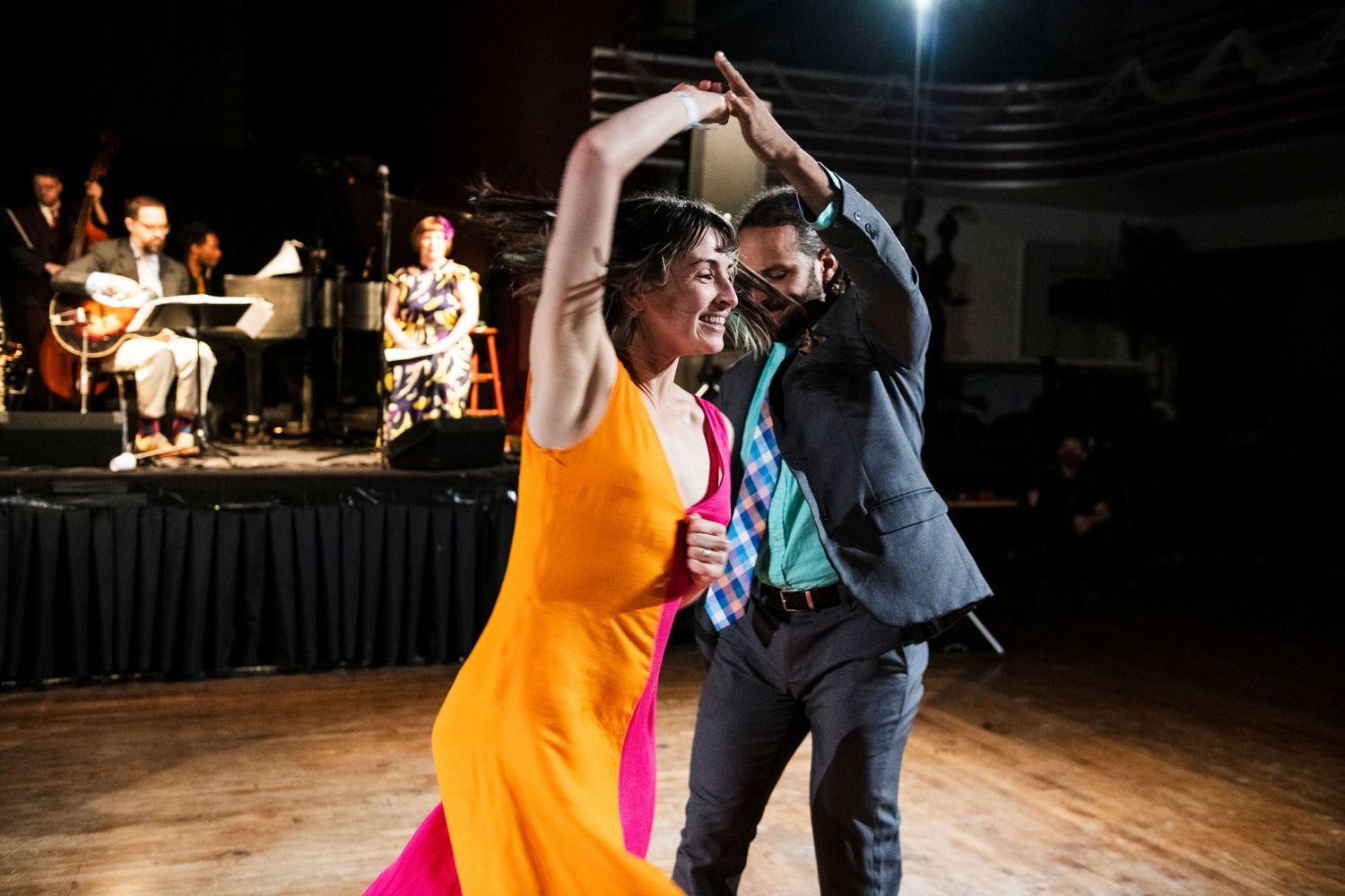 Dancers perform an energetic swing dance move on a wooden dance floor with a live band in the background.