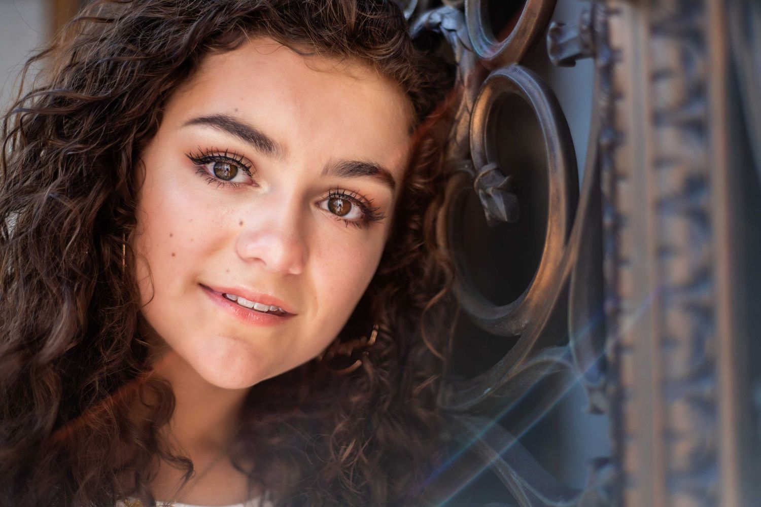 Portrait of someone with curly dark hair and natural makeup against an ornate iron background.