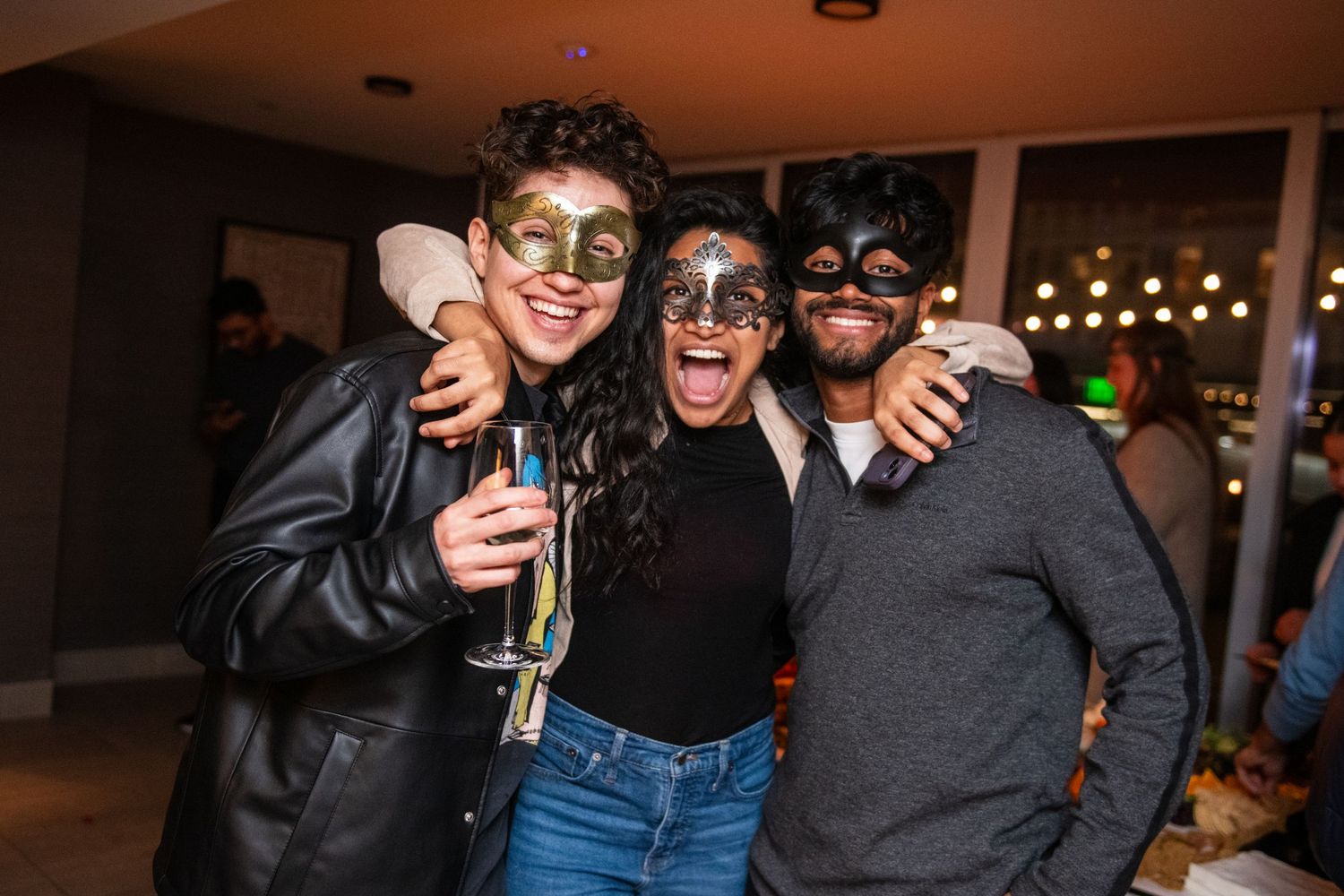 Friends in costume masks celebrate at an indoor party with drinks and laughter.