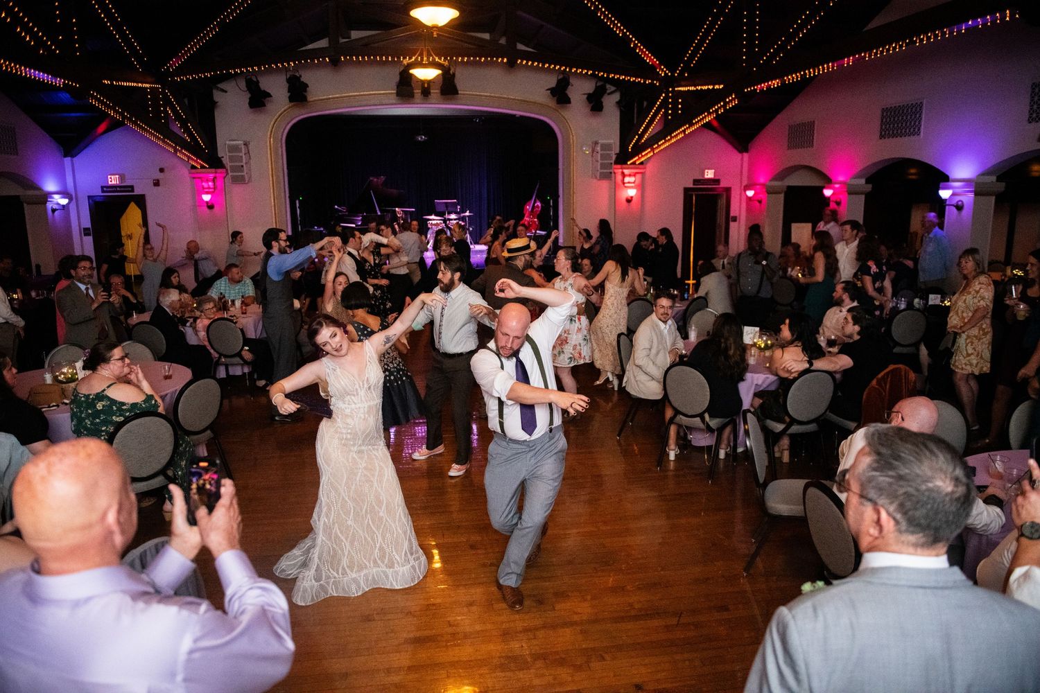Wedding celebration with guests dancing and celebrating on polished hardwood floors under purple lights.