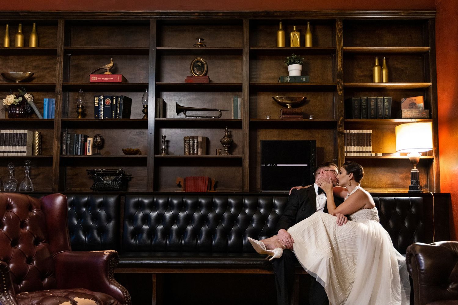 A couple relaxes on a leather couch surrounded by library bookshelves.