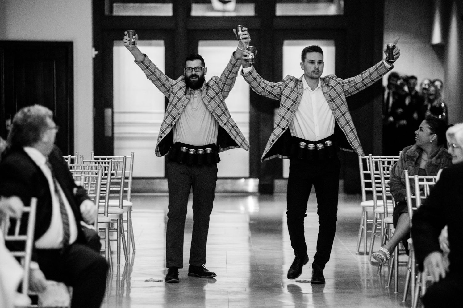 Two groomsmen joyfully walking down the aisle with raised arms at a wedding ceremony.