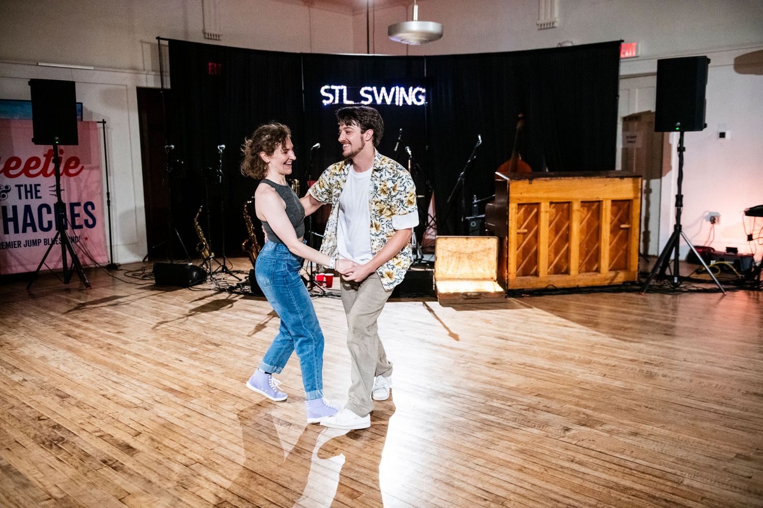 Swing dancers execute a partnered dance move on a hardwood floor with STL Swing banner behind.