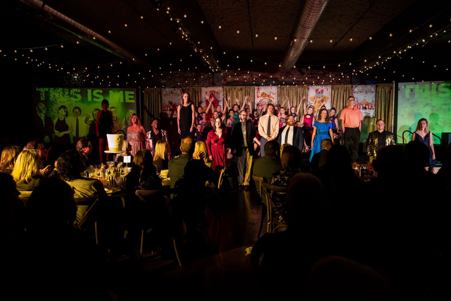 Group performance on stage with colorful lighting and audience silhouettes in foreground.