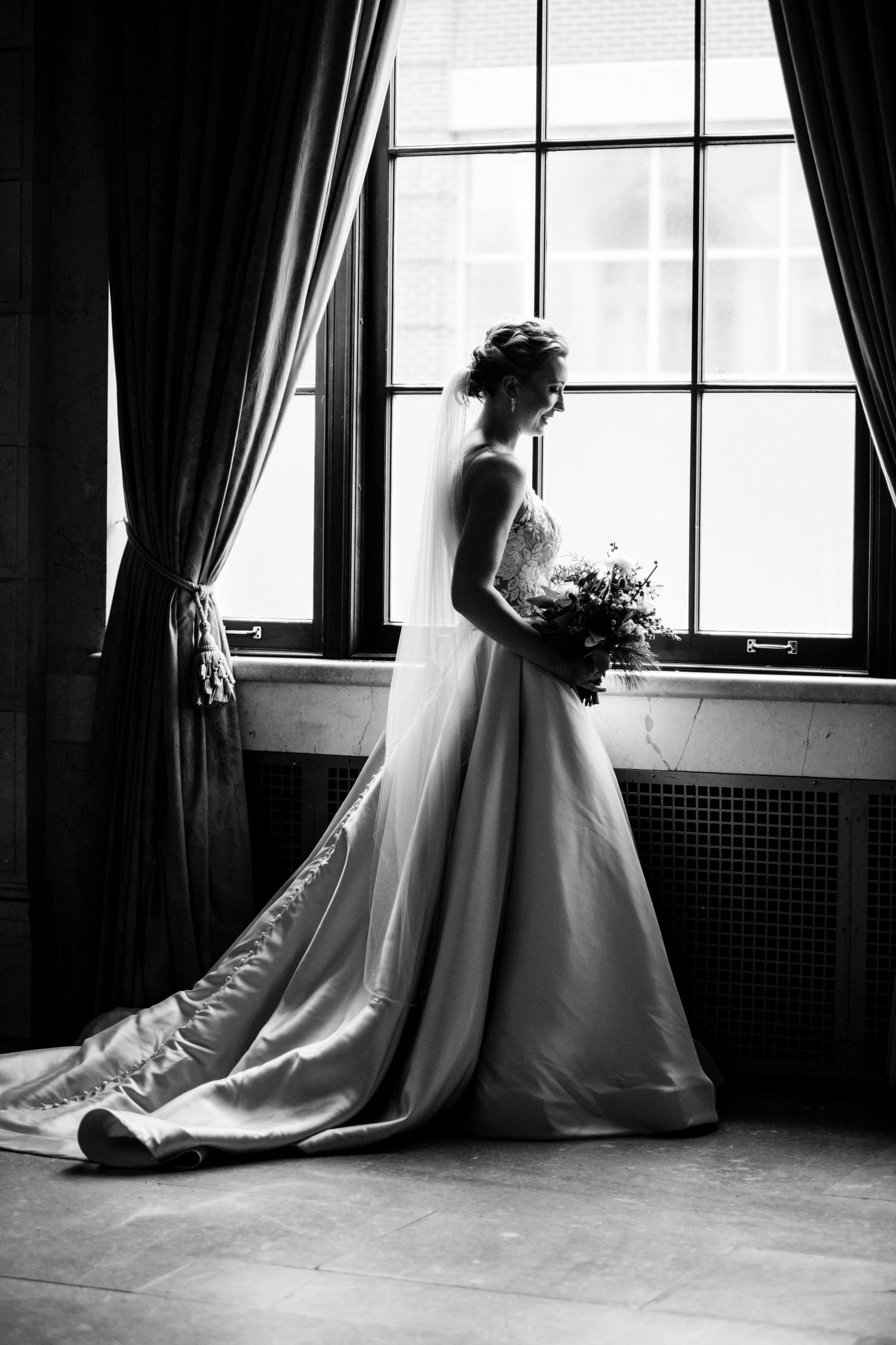 Silhouetted bride poses by window holding bouquet in flowing white gown and veil.