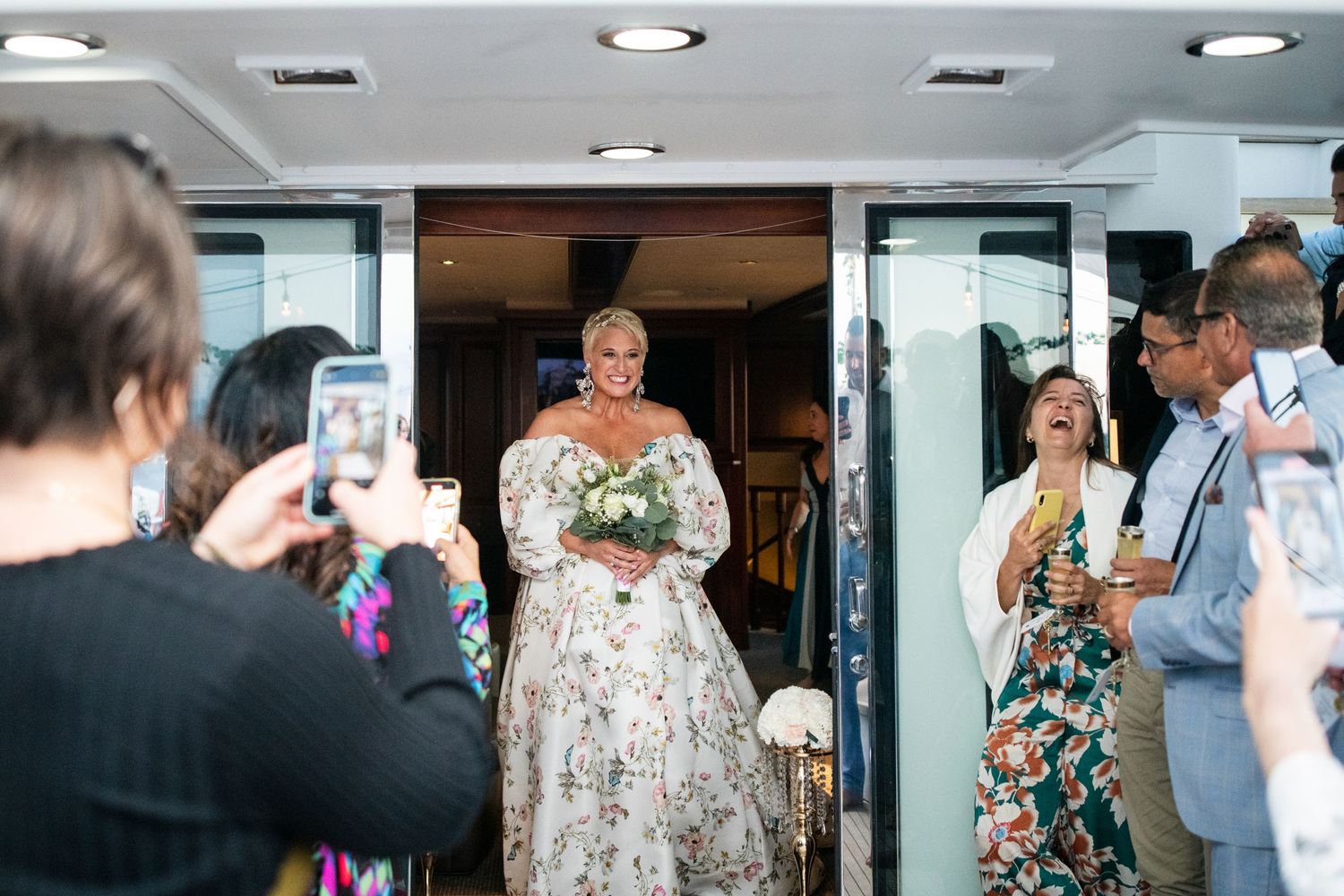 Wedding guests take photos as bride in floral dress makes grand entrance.