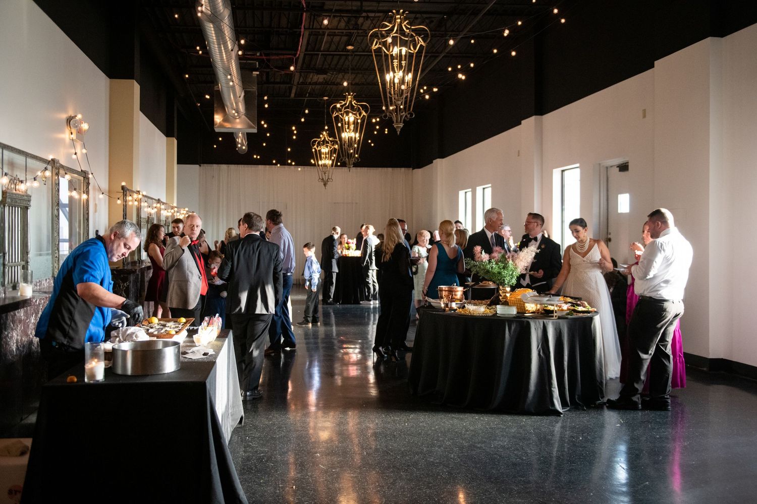 Event guests mingling around buffet tables in an elegant venue with hanging lanterns and twinkle lights.