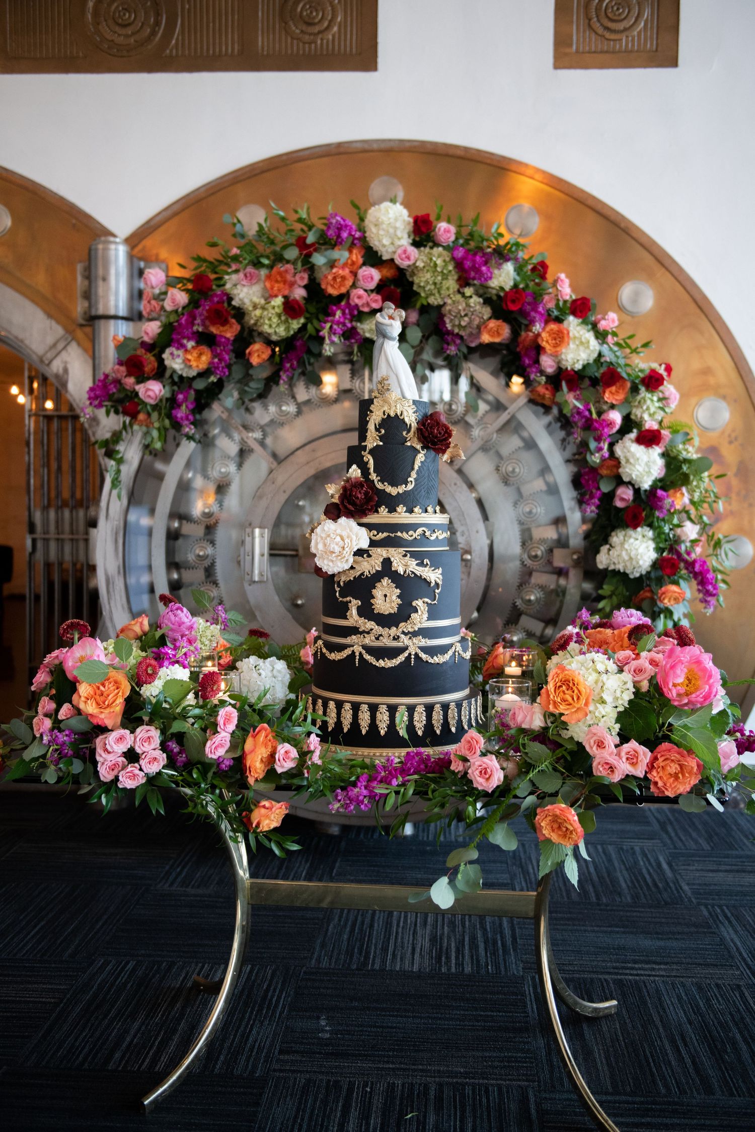 Elegant black wedding cake with gold details displayed under a floral arch with pink and purple blooms.