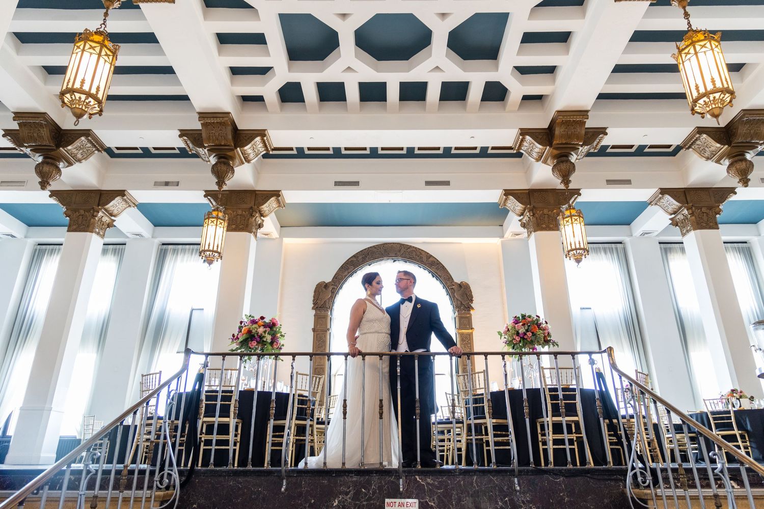 A couple stands together on an ornate balcony with decorative columns and lighting fixtures.