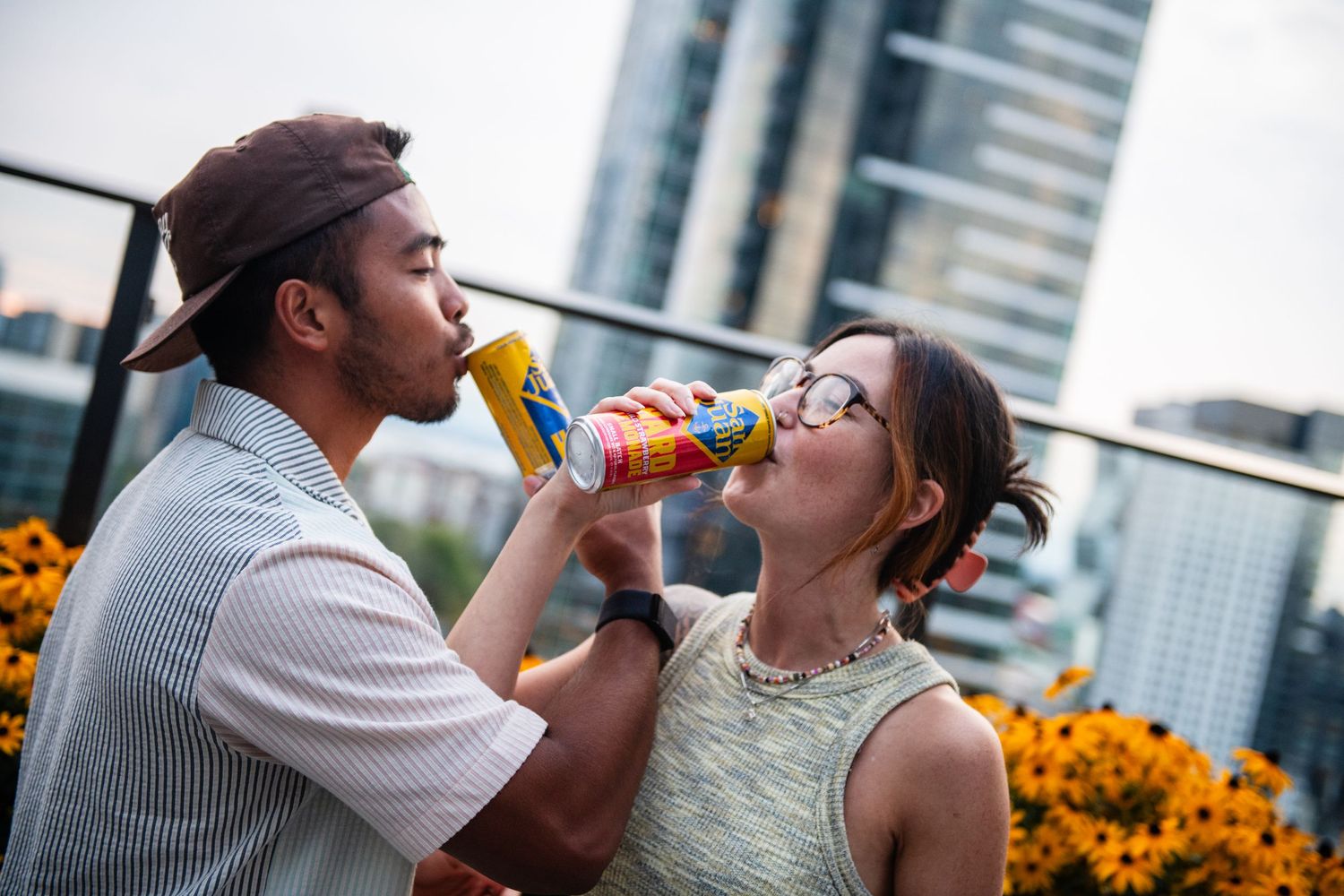 Couple sharing drinks on a rooftop terrace with yellow flowers and skyscrapers.