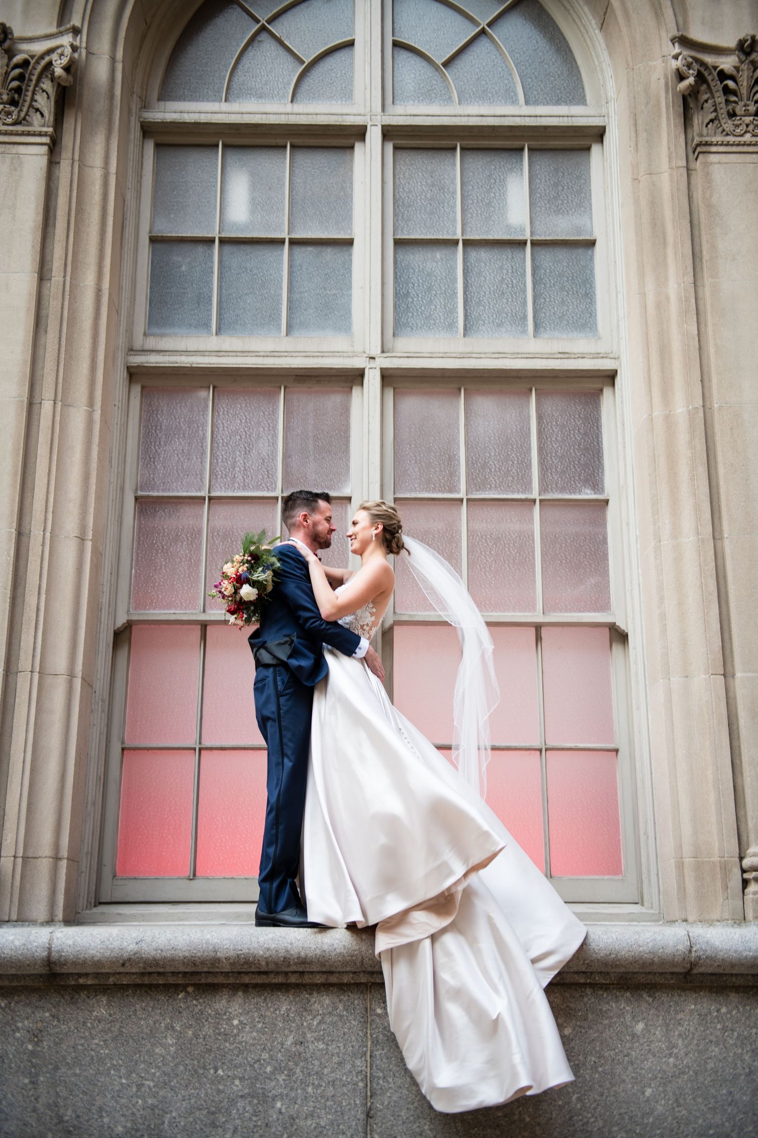 Bride and groom share romantic moment against grand arched window with pink glow.