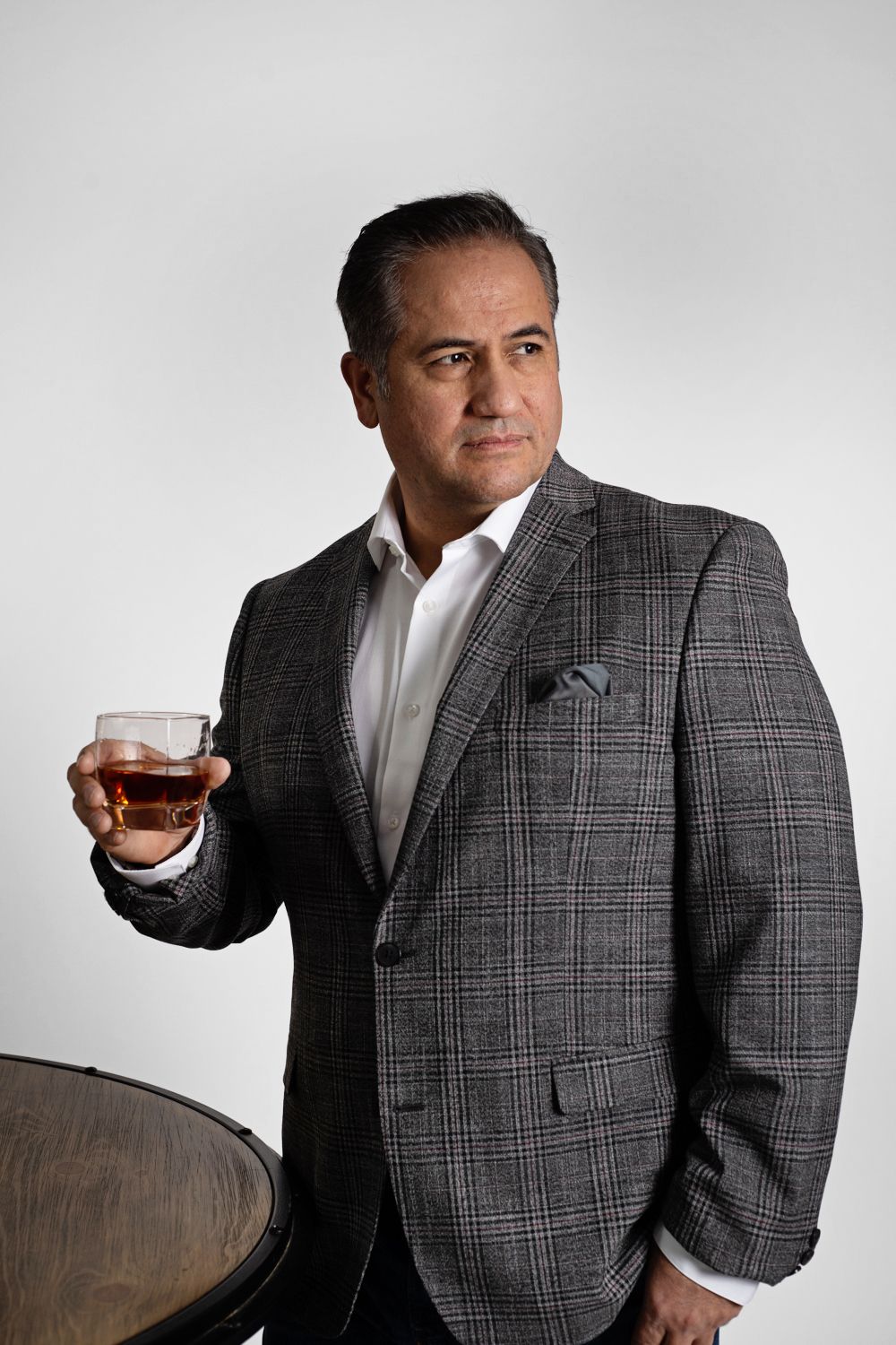 Well-dressed man in grey plaid suit jacket holding whiskey glass against white backdrop.