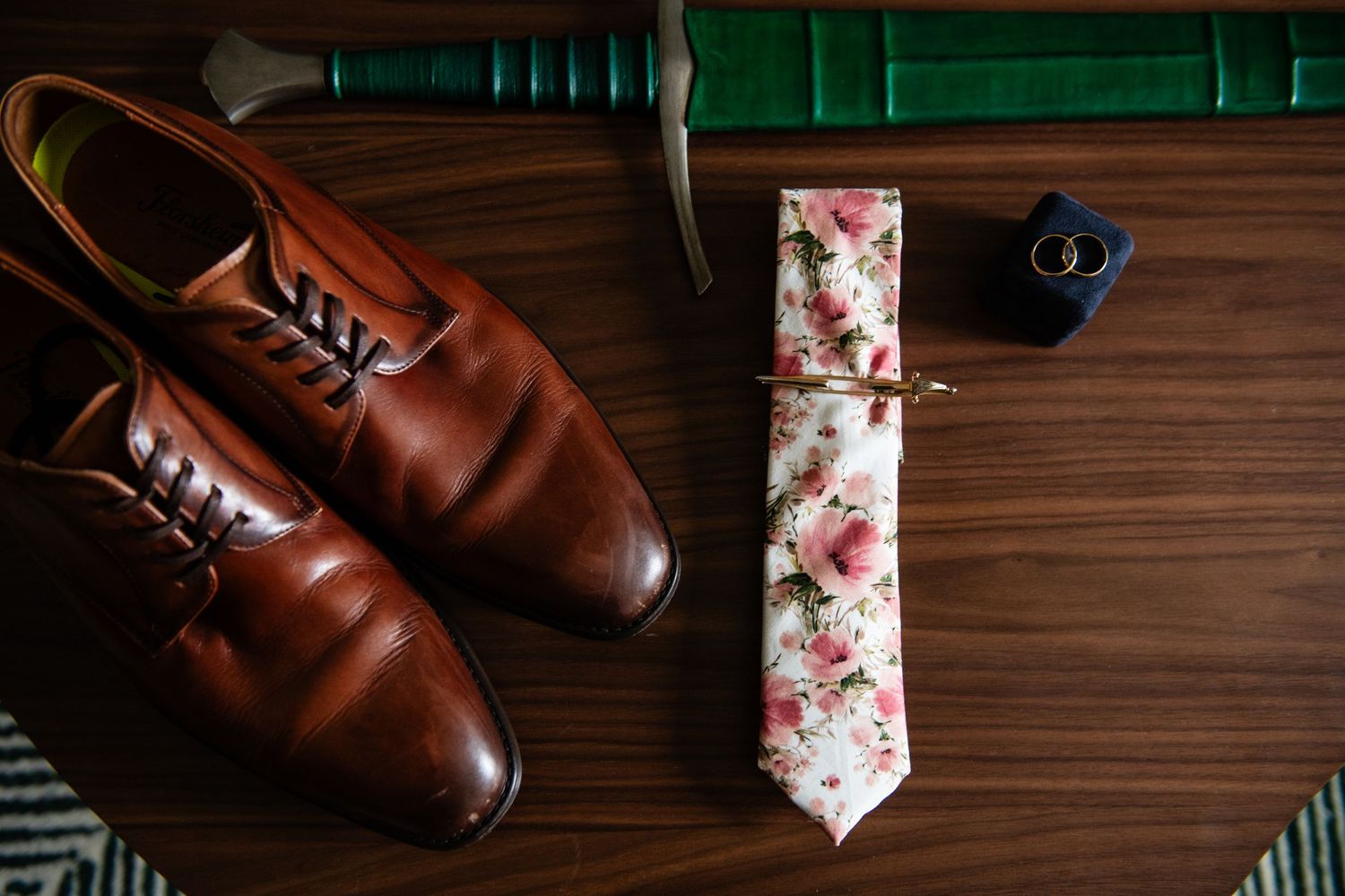 Brown leather dress shoes, floral tie, and ring box arranged on wooden surface for wedding details.