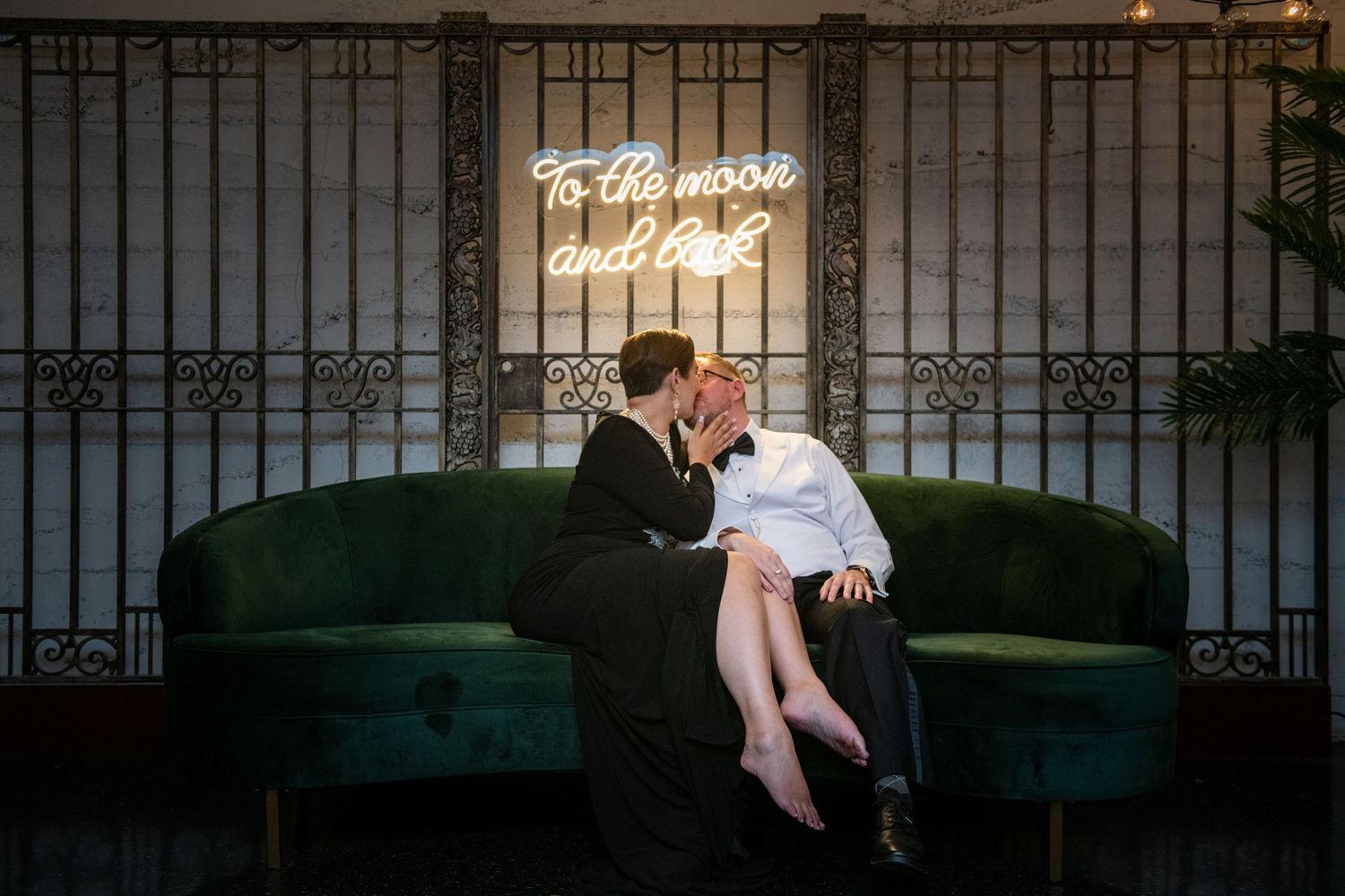 A couple shares an intimate moment on a green velvet couch beneath neon lighting.