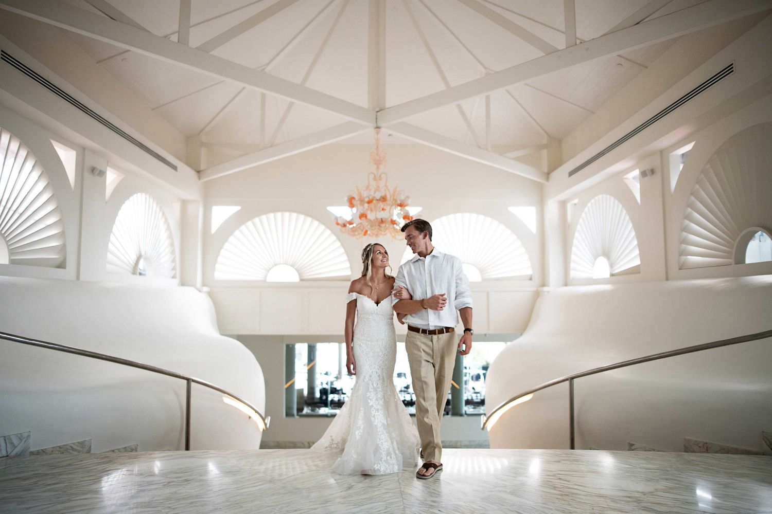 A wedding couple descends elegant white stairs in a bright architectural setting with arched windows.