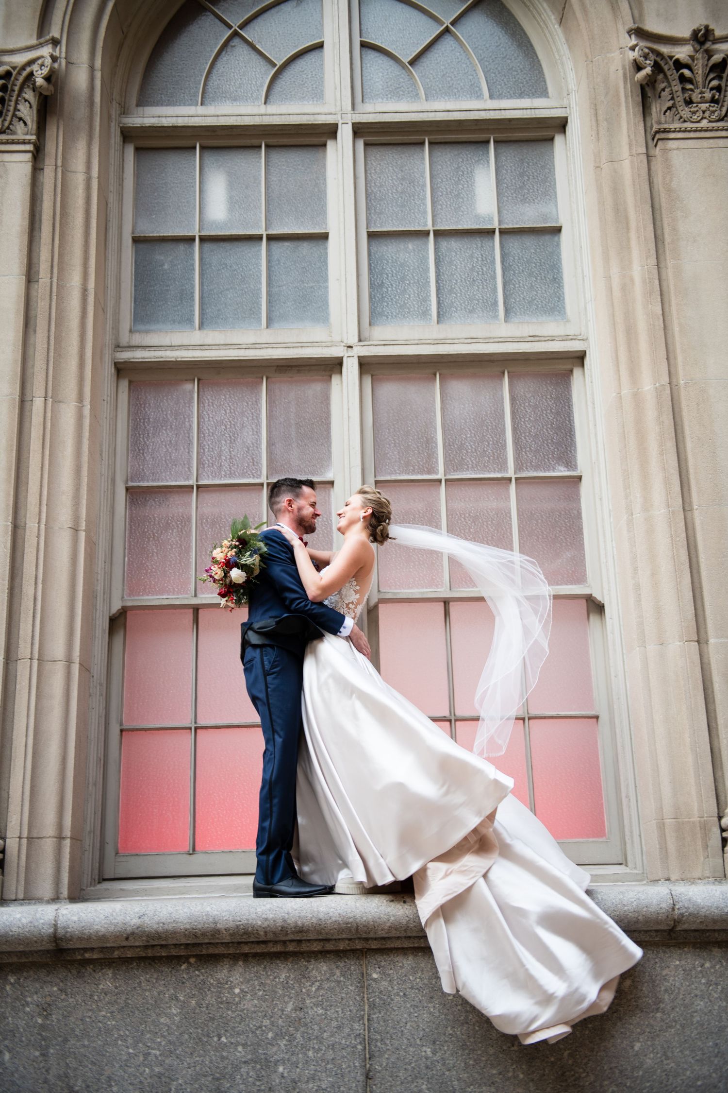 Newlyweds share intimate moment against pink-tinted window with flowing wedding dress.