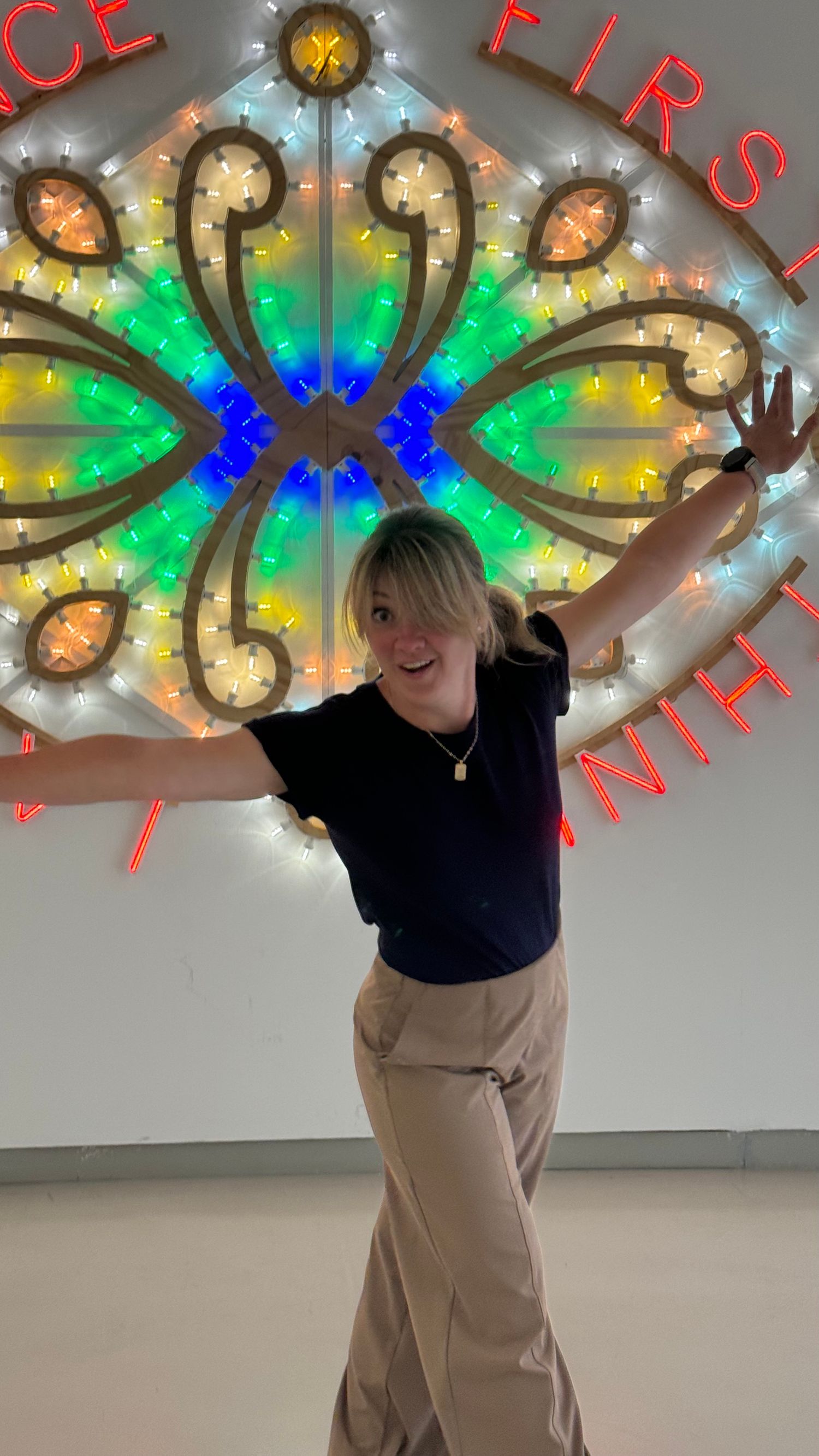 Person posing joyfully under colorful illuminated art installation with flower-like design.