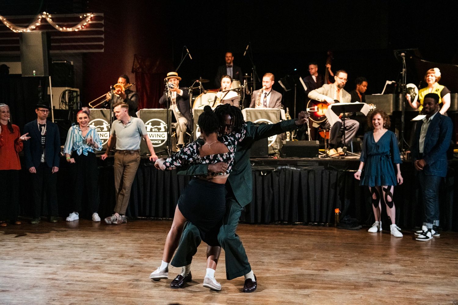 Swing dancers perform on a wooden dance floor in front of a big band orchestra on stage.