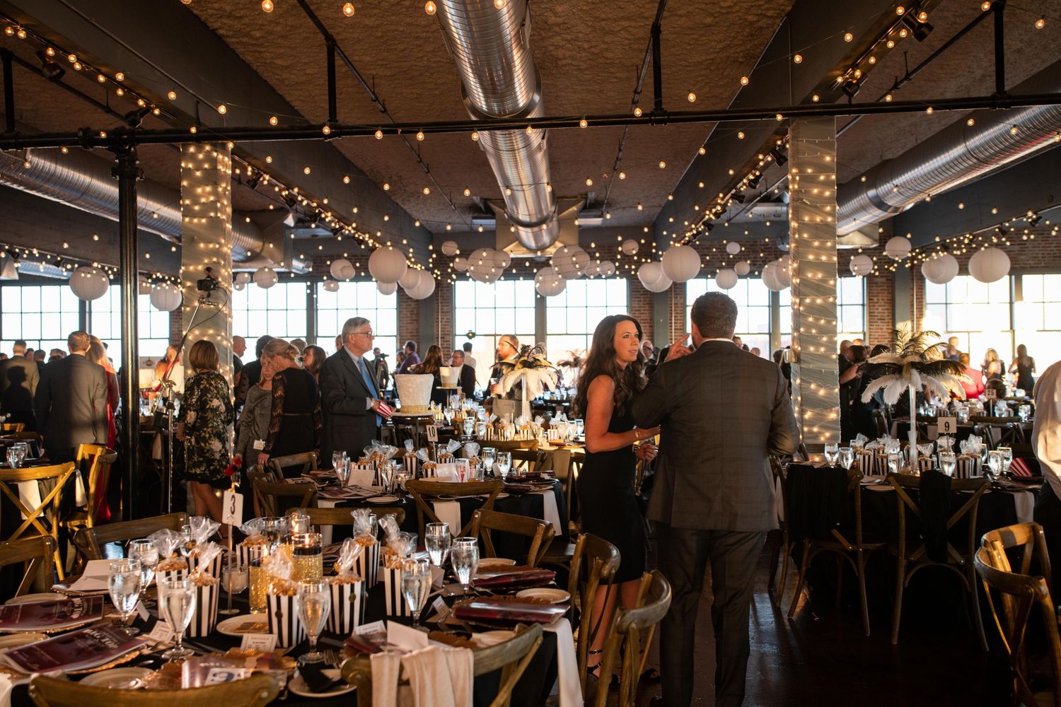 Rustic event space with exposed brick walls and string lights as attendees gather at formal dinner tables.