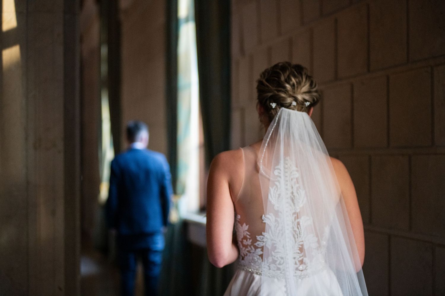 Artistic shot of bride in white lace gown with veil from behind looking towards silhouette.