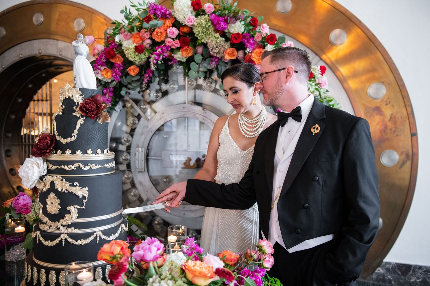 Elegant black wedding cake decorated with colorful floral arrangements against an illuminated vault door.