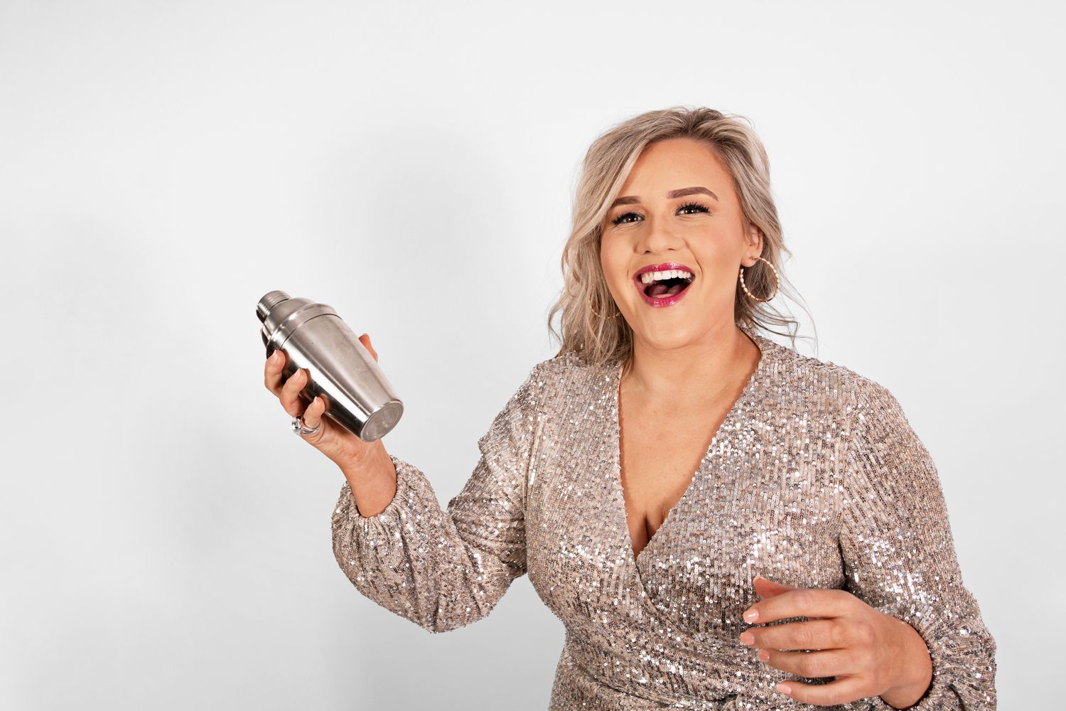 Person in sparkly dress laughing while holding cocktail shaker against white background.