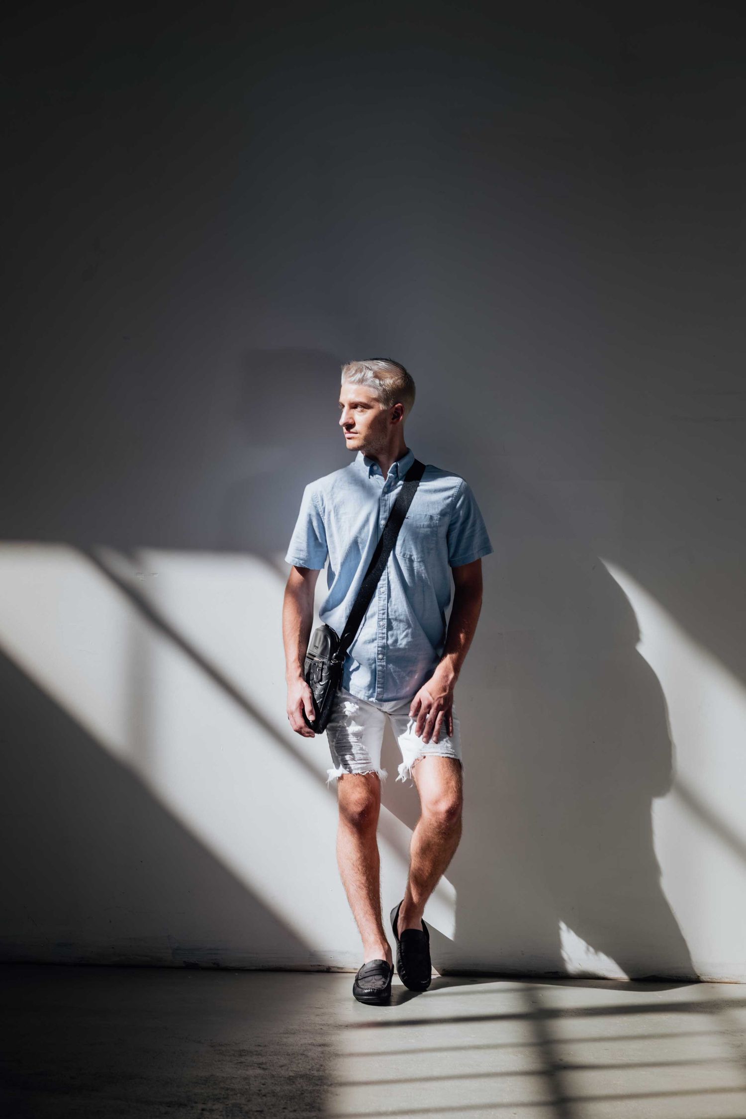Person in casual summer attire standing against white wall with dramatic diagonal shadows and lighting.