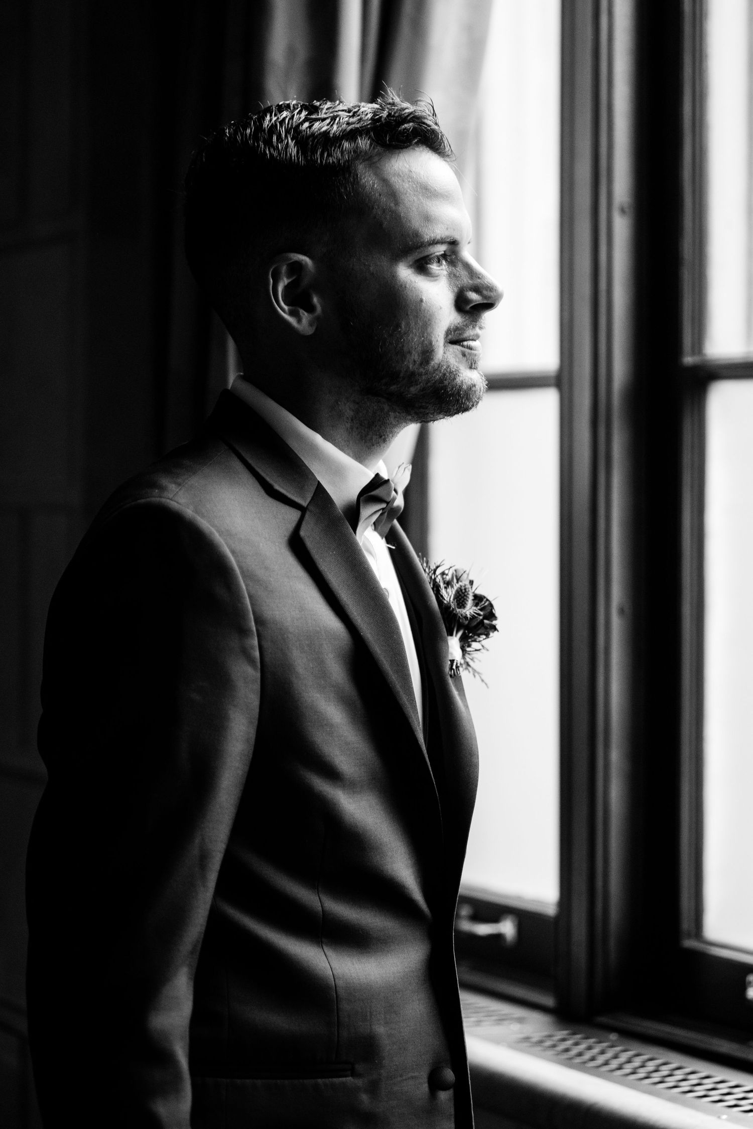 Side profile of groom in suit gazing thoughtfully out window in black and white.