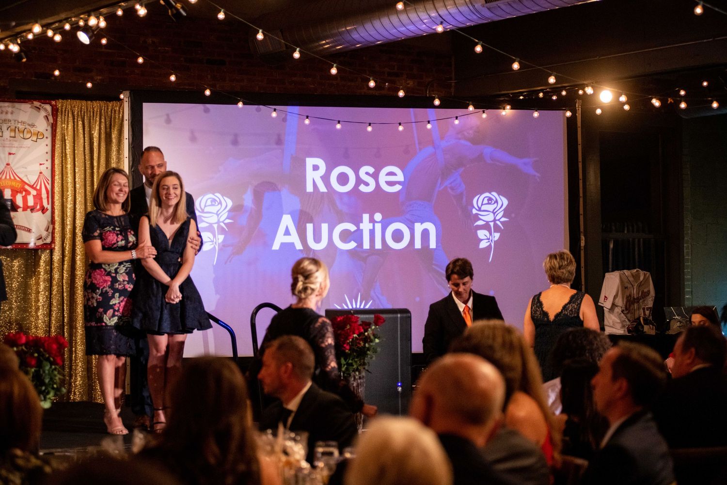 Rose Auction event stage with string lights and purple backdrop during a formal gathering.