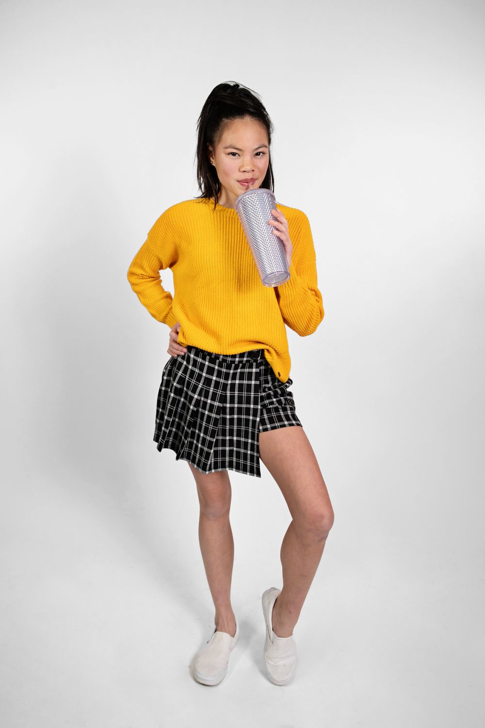 Young woman in bright yellow sweater and plaid skirt posing with silver tumbler against white background.