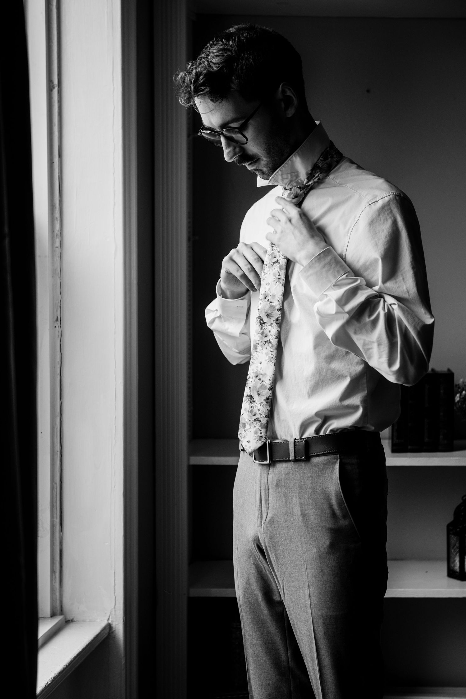 A black and white portrait of someone tying their necktie by a window.