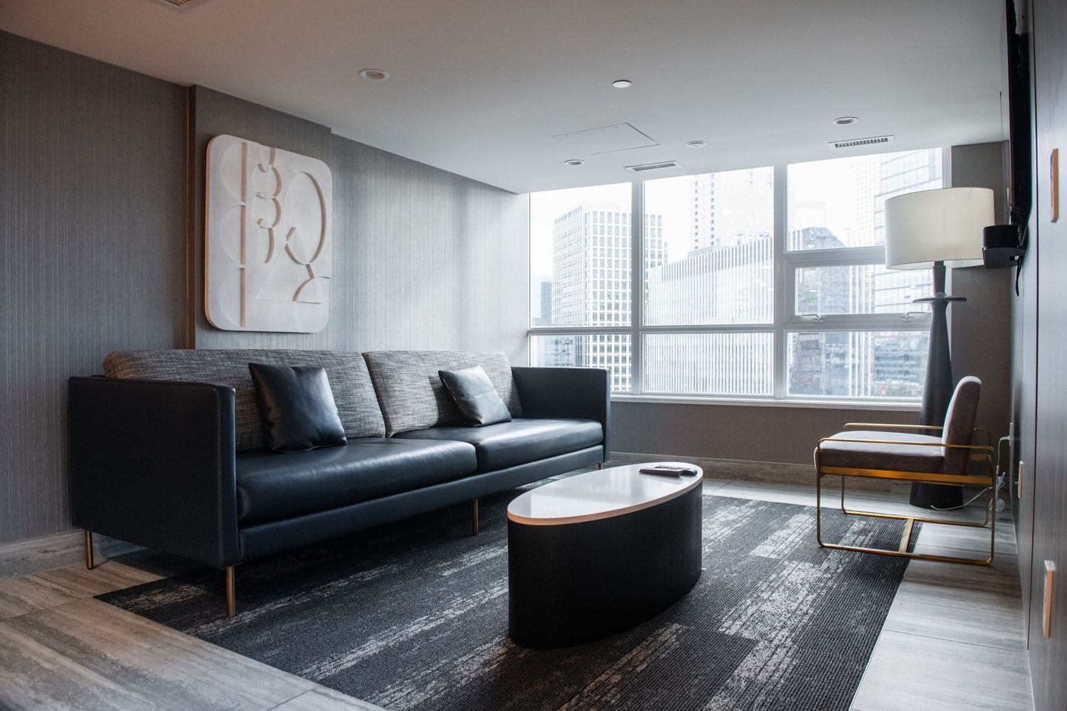 Modern living room with navy leather sofa, oval coffee table, and large windows overlooking city buildings.