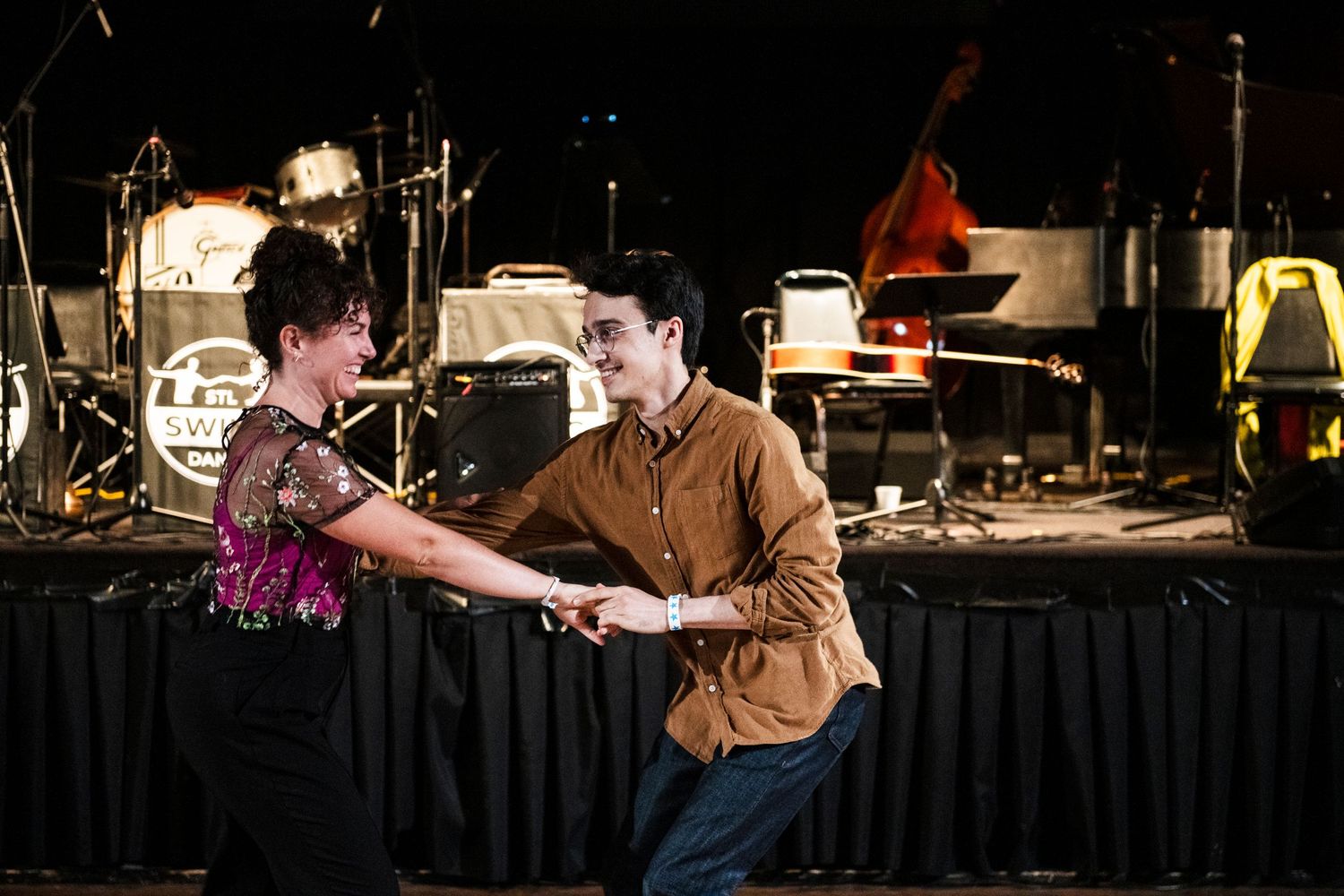 Two dancers practice swing dance moves in front of a stage with musical instruments.