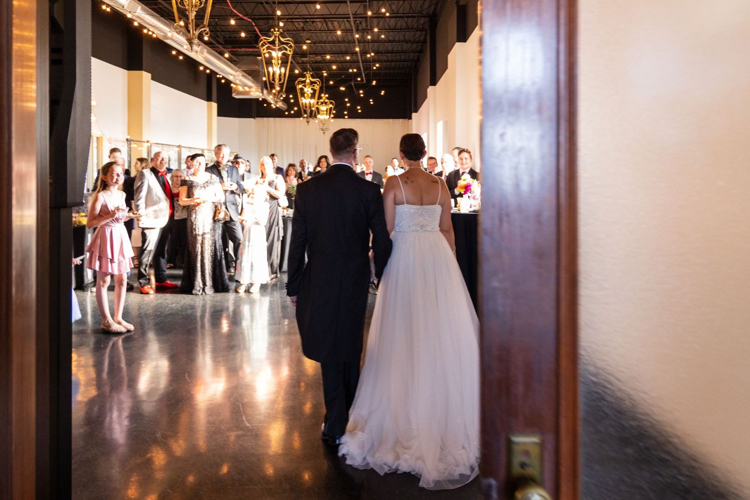 Wedding couple walking down hallway with string lights as guests line the corridor.