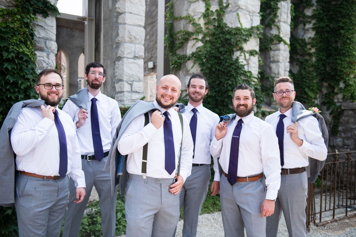 Groomsmen in matching gray suits and purple ties pose together against stone wall with ivy.