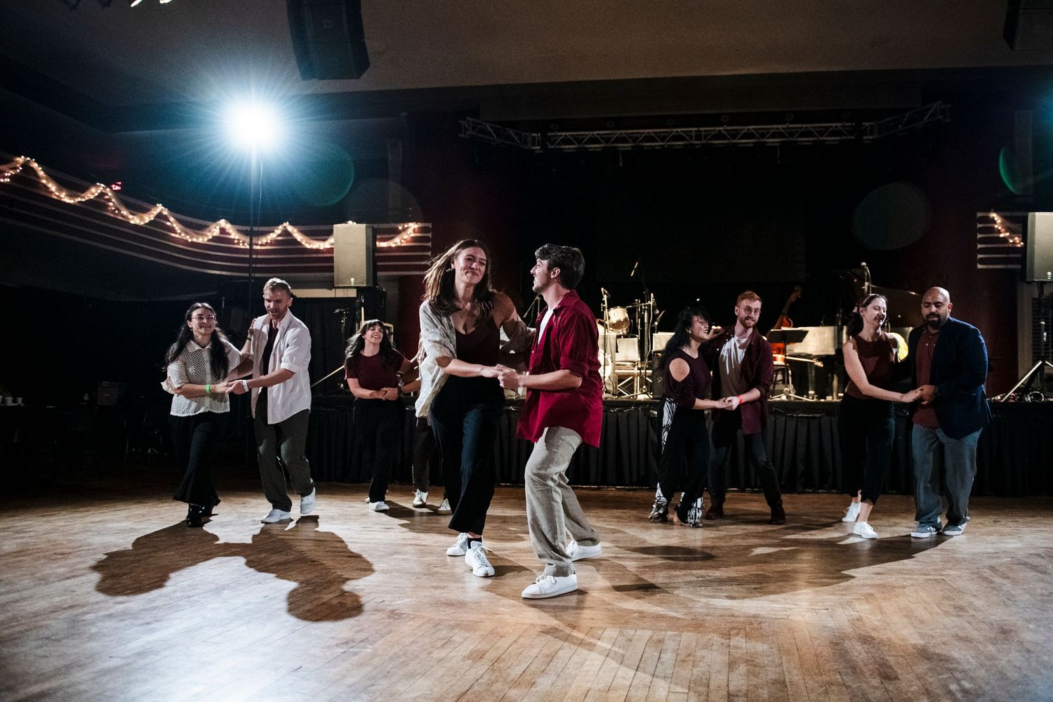 Swing dance performers practice their routine on a polished wooden floor with band setup visible.