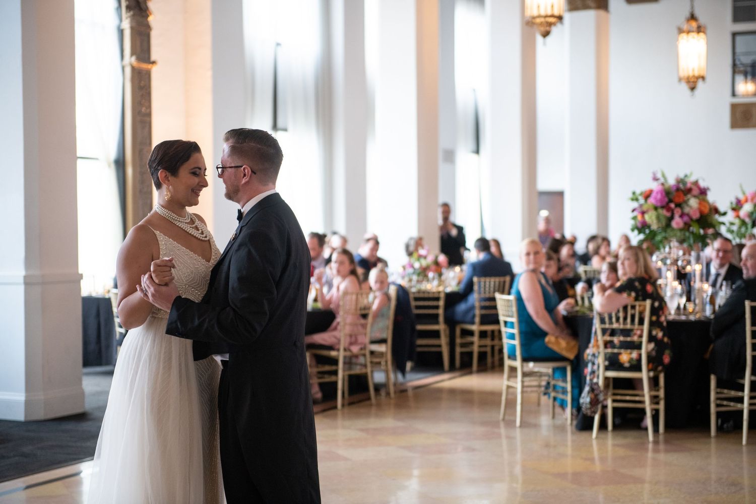 Intimate dance moment in elegant ballroom with ornate columns and dining tables in background.