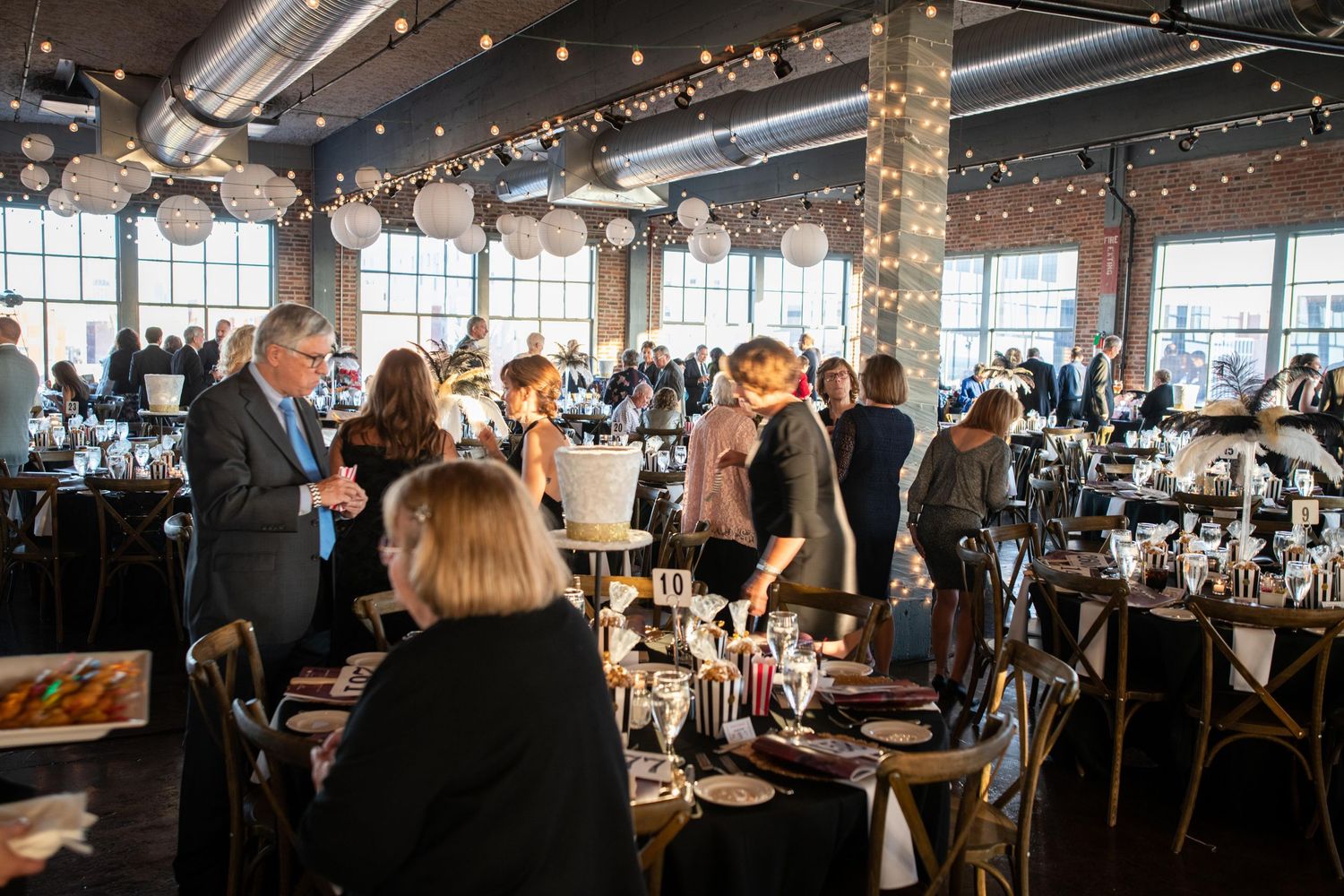Elegant event space with string lights and large windows where guests mingle at decorated tables for a formal gathering.