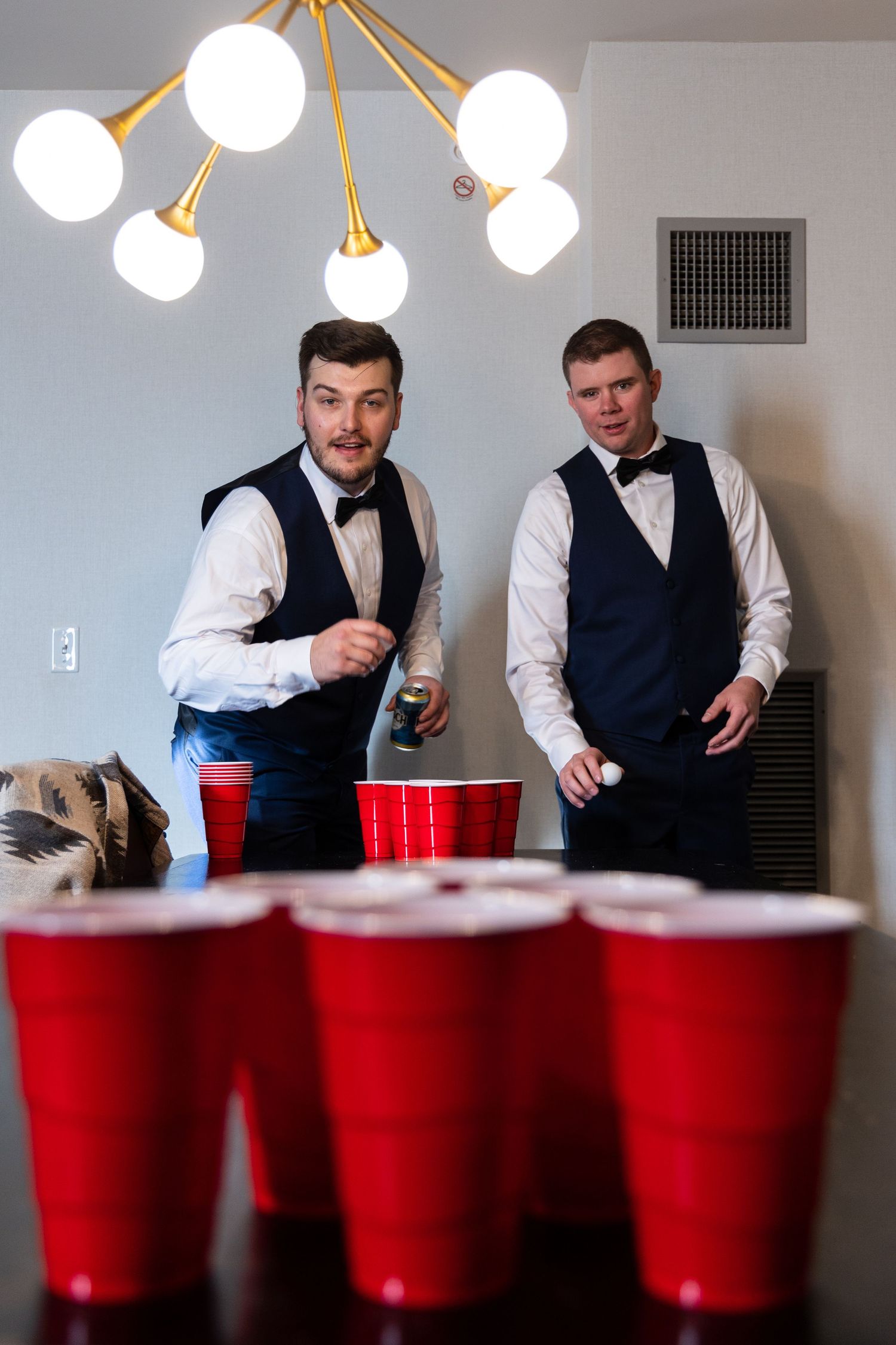Two groomsmen in formal attire playing beer pong with red cups under a modern light fixture.