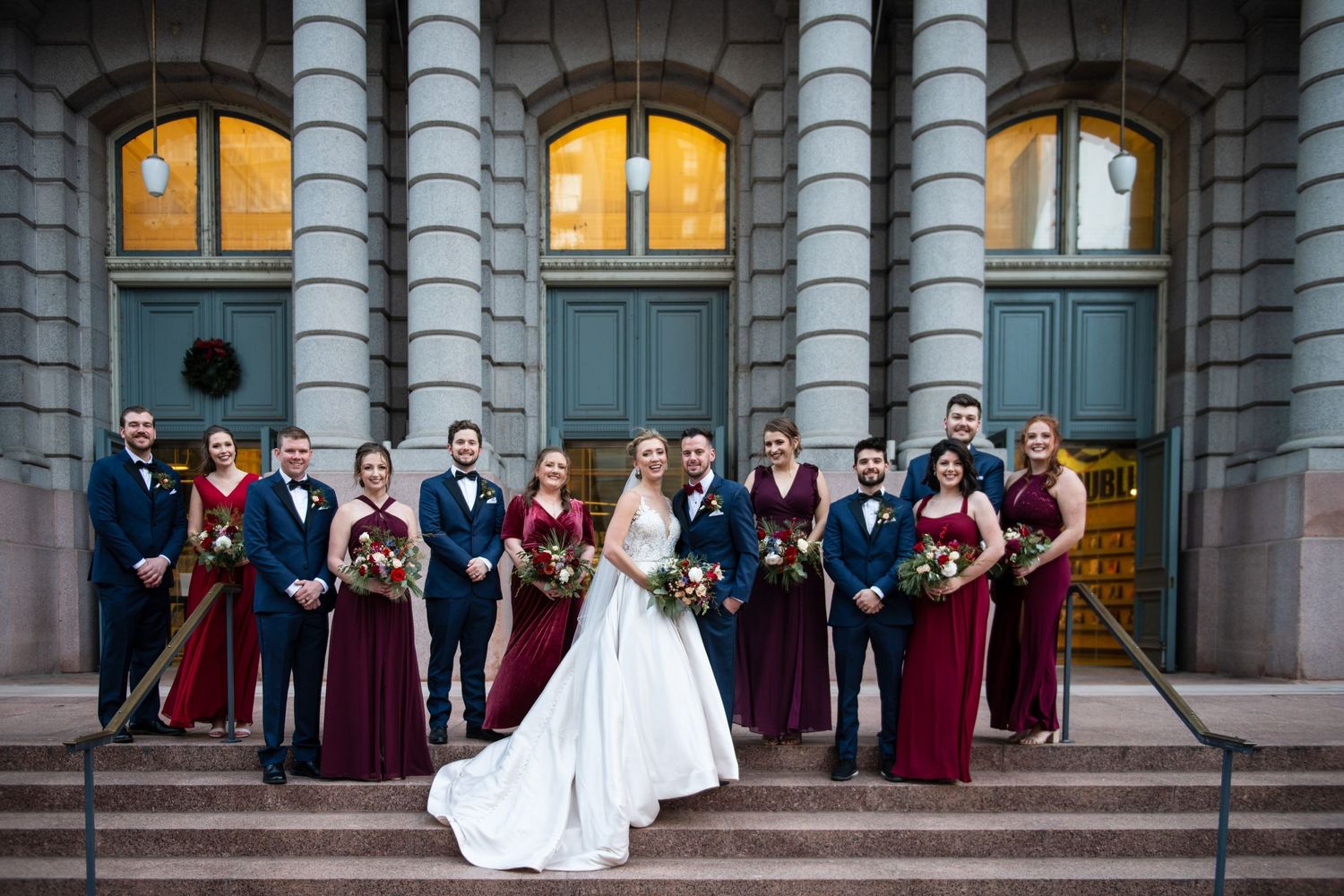 Wedding party poses on stone steps of historic building with burgundy bridesmaids dresses and navy suits at dusk.