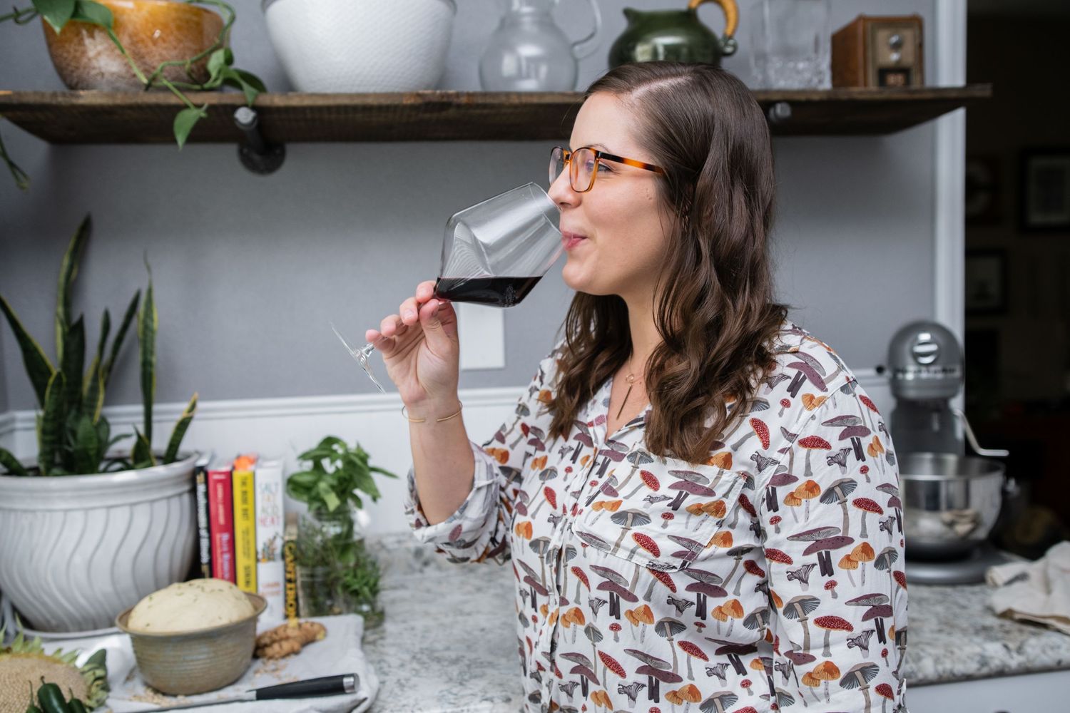 A person sipping red wine while standing in a kitchen with rustic wooden shelves.