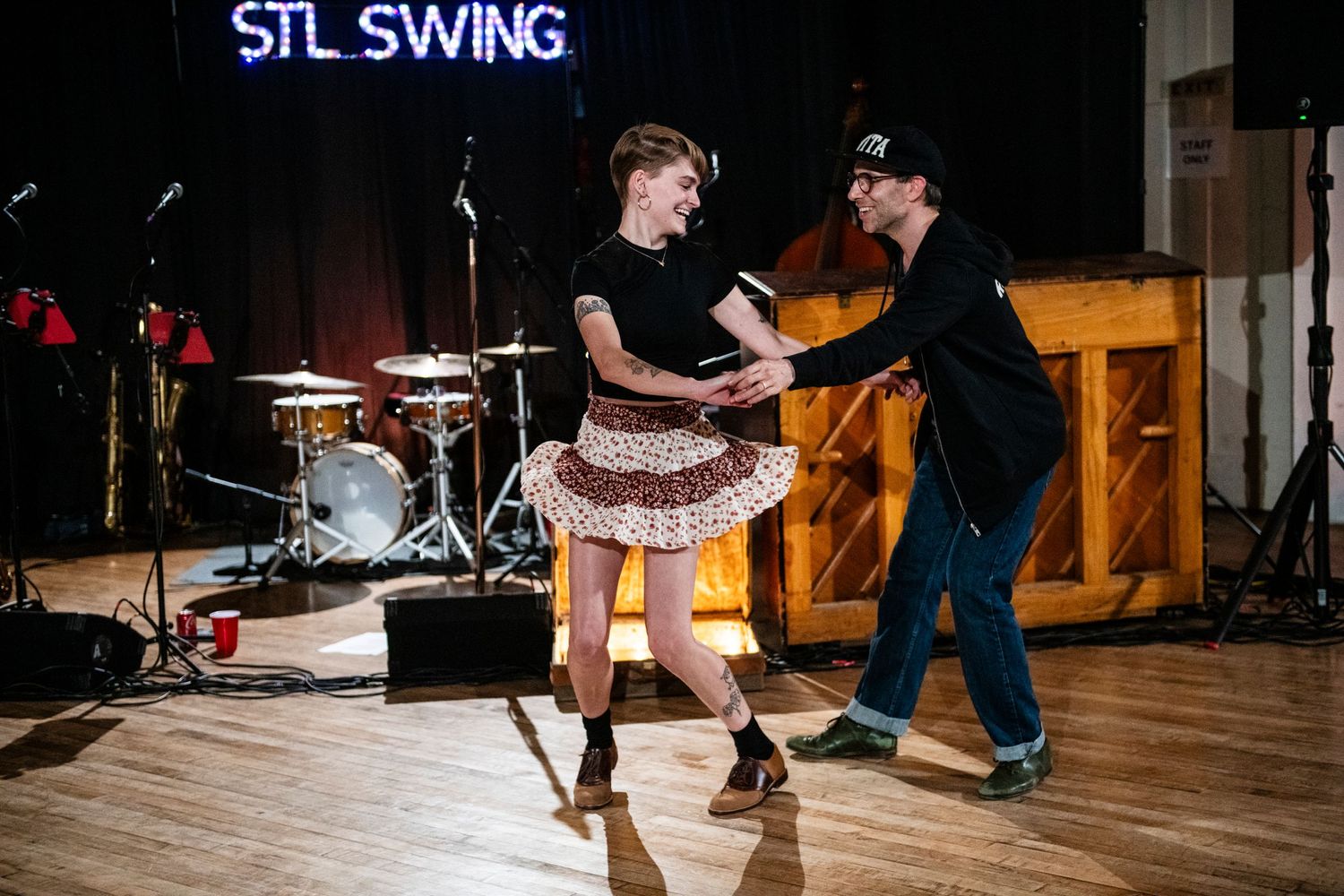 Dancers perform vintage-style swing moves on wooden dance floor with drum kit visible.
