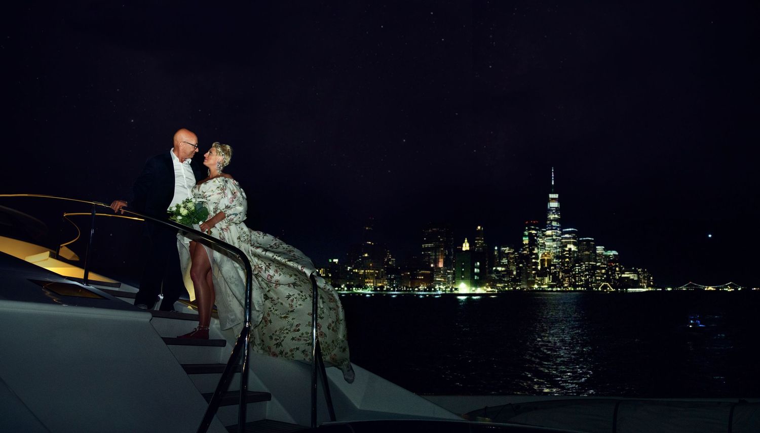 Couple stands at yacht railing with illuminated Manhattan skyline visible across the water.