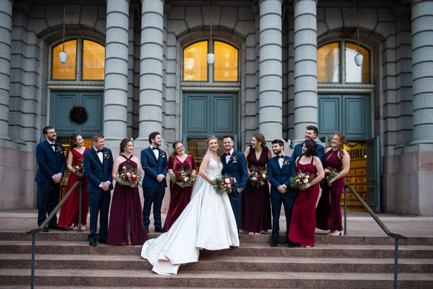 Elegant wedding party stands on architectural steps with warm lighting in background.