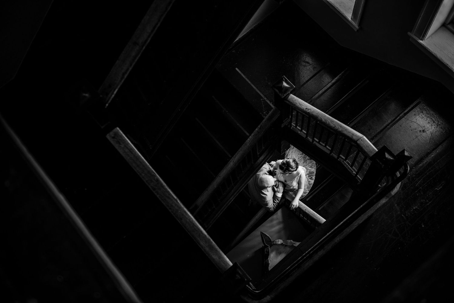 A dramatic black and white aerial shot of a couple on a dark spiral staircase.